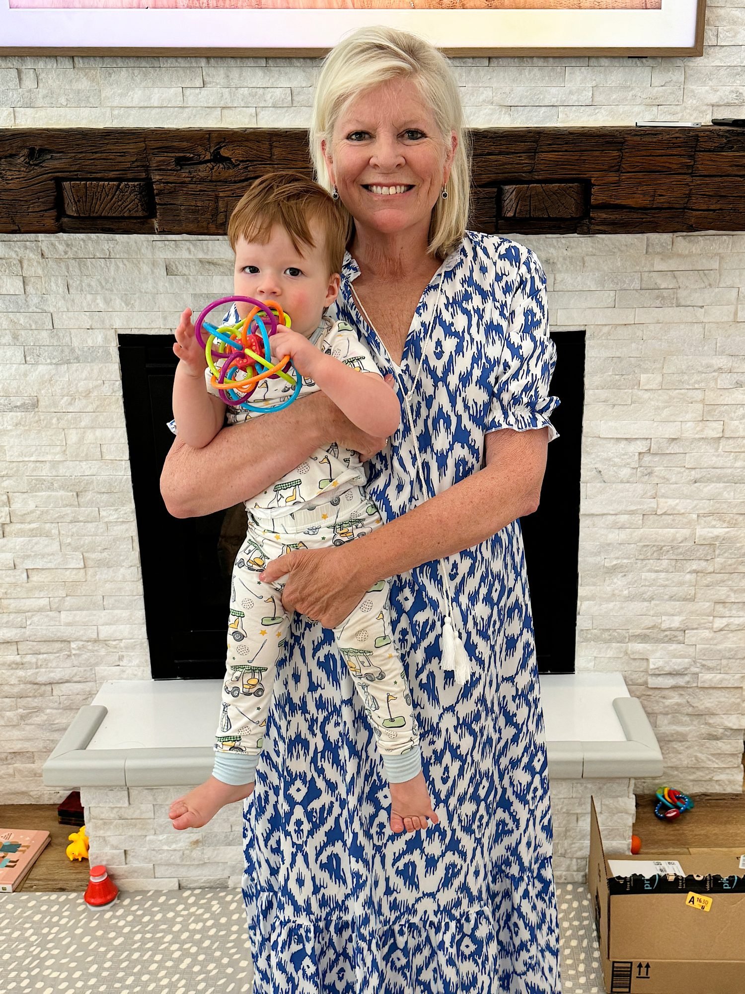 A smiling woman in a blue patterned dress holds a toddler who is holding a colorful toy against a brick fireplace with a wooden mantle.