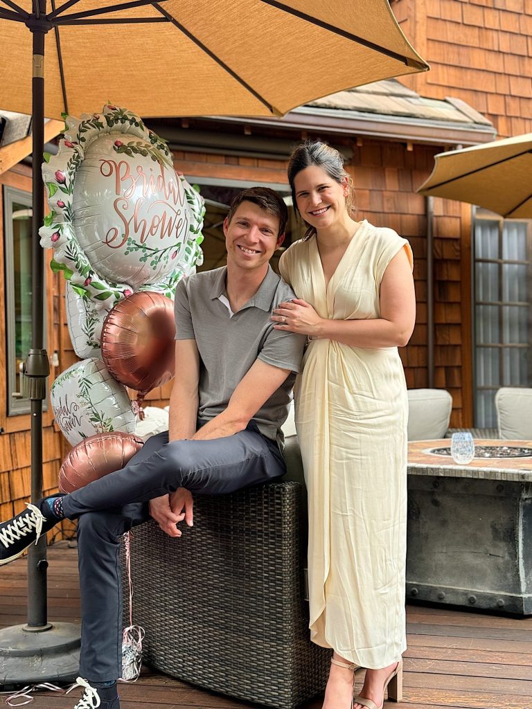 A smiling man and woman pose on a patio next to a "Bridal Shower" balloon arrangement, under large patio umbrellas. The man is seated while the woman stands, resting her hand on his shoulder.