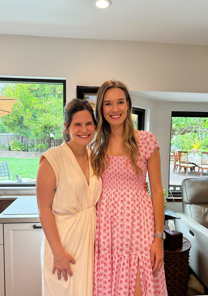 Two women standing indoors, smiling. The woman on the left wears a white dress, and the woman on the right wears a pink floral dress. They are in a room with large windows showing a garden outside.