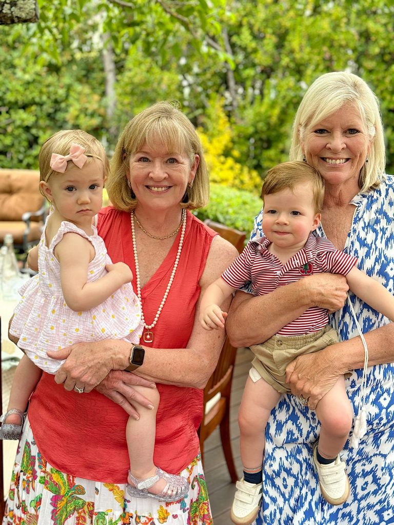 Two women hold two toddlers outdoors in a garden with greenery in the background. One woman wears a red top and skirt, the other a blue and white dress. The toddlers wear summer clothes.