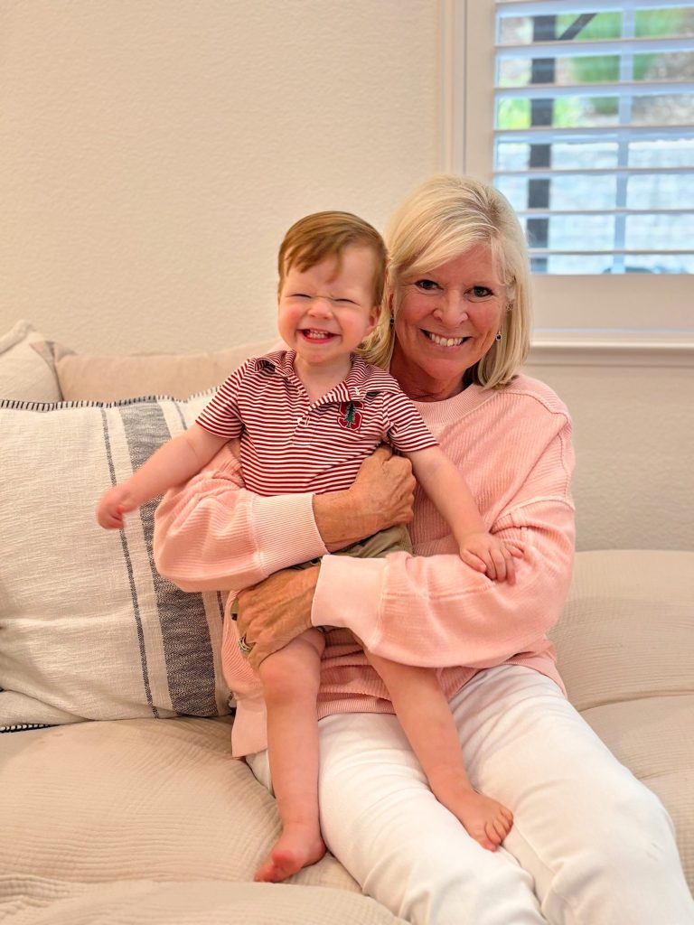 A woman wearing a pink sweater is sitting on a sofa, holding a smiling baby dressed in a red and white striped shirt and beige shorts. There's a window with blinds in the background.