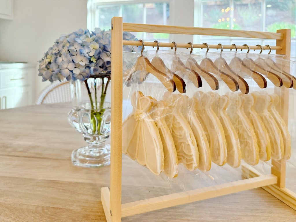 Cookies shaped like dresses hang on mini wooden hangers, displayed on a stand next to a vase of blue hydrangeas on a wooden table.