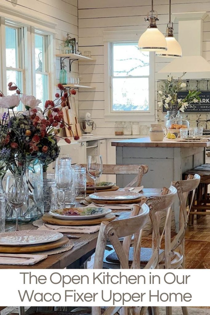 A rustic kitchen with wooden furniture, a dining table set for a meal, and various kitchen items displayed. Shelving holds decor and kitchenware. Two pendant lights hang over the kitchen island.