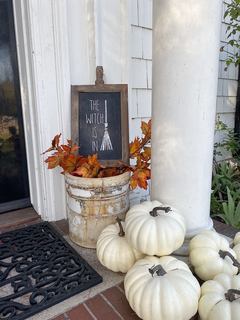 A chalkboard sign reading "The Witch is In" with a broom illustration stands in a white pot with orange leaves, placed next to white pumpkins and a column by a front door.