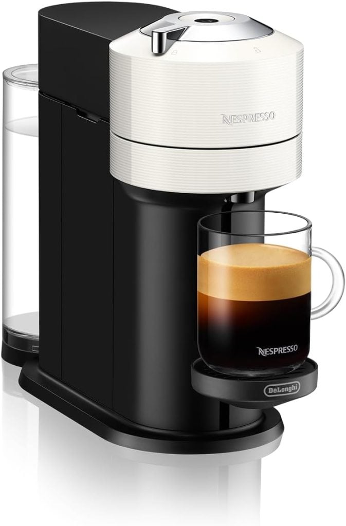 A sleek black and white Nespresso coffee machine with a glass mug placed on the drip tray, half-filled with coffee. The machine features a water reservoir on the back.