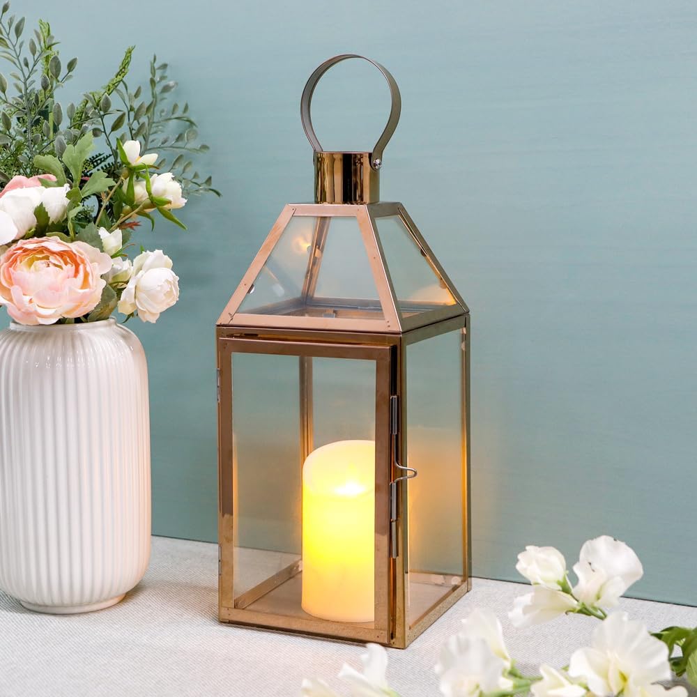 A lit candle inside a glass lantern sits on a table next to a white vase containing pink and white flowers arranged in a delicate fall flower arrangement against a light blue background.