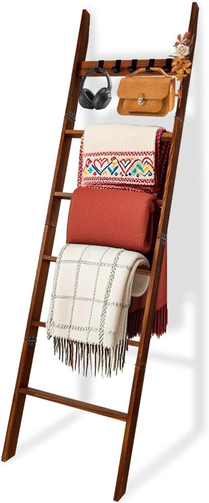 A wooden ladder-style rack holds a pair of headphones, a brown bag, a flower, a folded embroidered blanket, a solid red blanket, a plaid blanket, and a small fall pumpkin for seasonal charm.