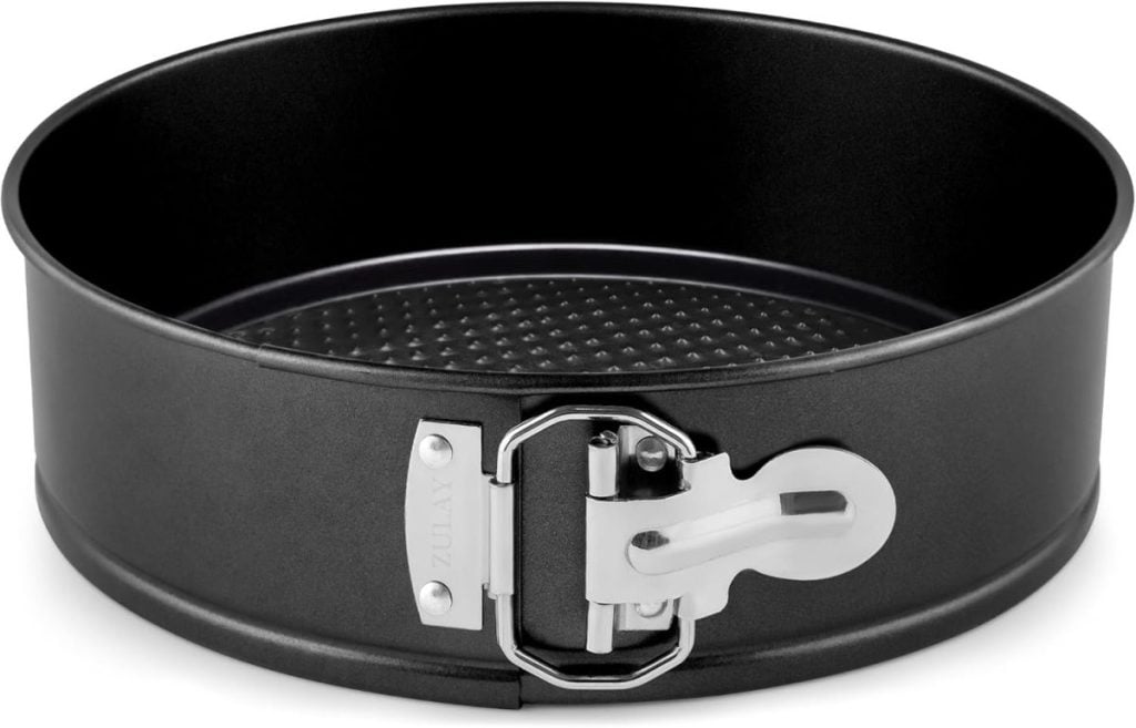 A round, black non-stick springform pan with a latch mechanism on the side. The brand name "Zulay" is engraved on the latch.