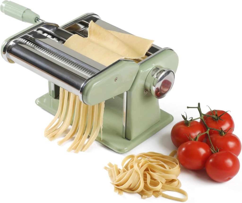 A manual pasta maker cuts fresh pasta dough into strips, perfect for your favorite Fall Pasta Recipes, with a small bunch of tomatoes beside the finished pasta on a white background.