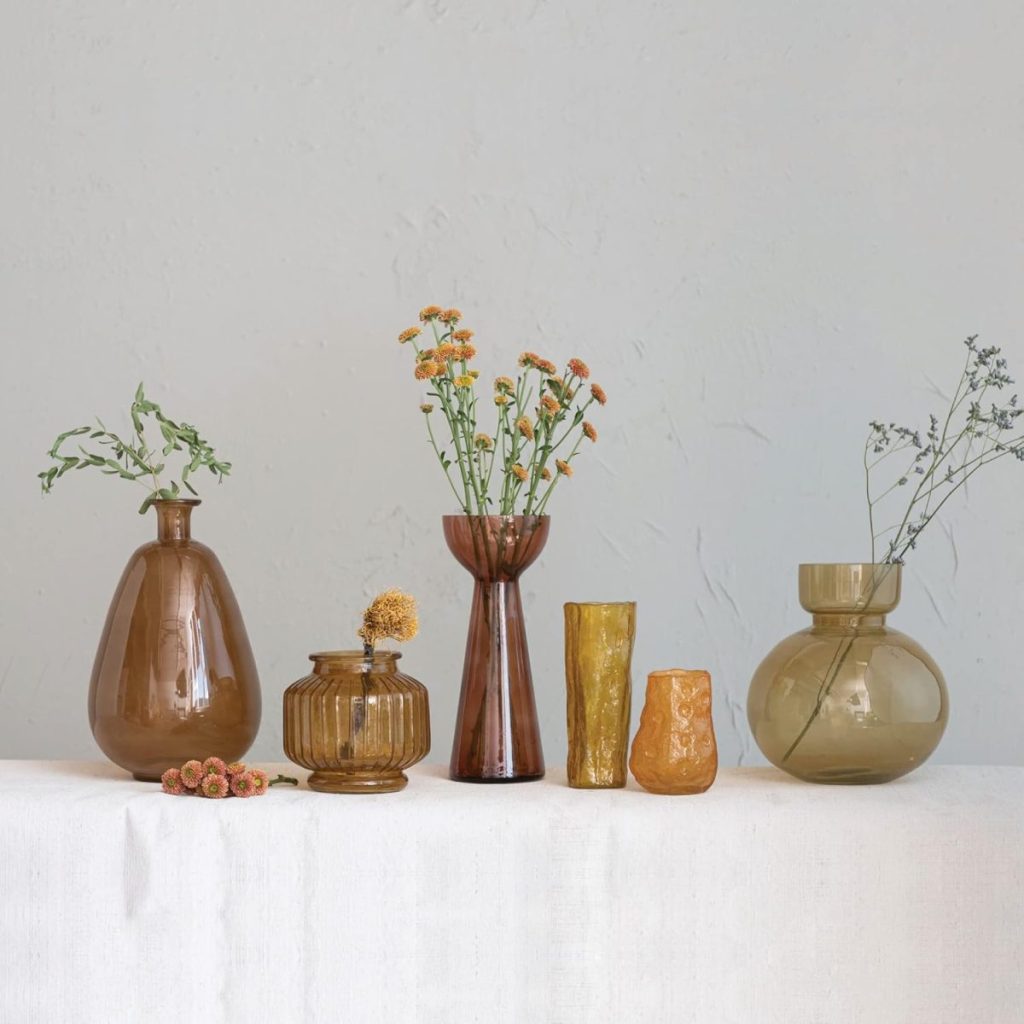 A row of amber and yellow glass vases with fall flower arrangements sits on a light-colored table, each filled with small seasonal blooms against a plain wall.