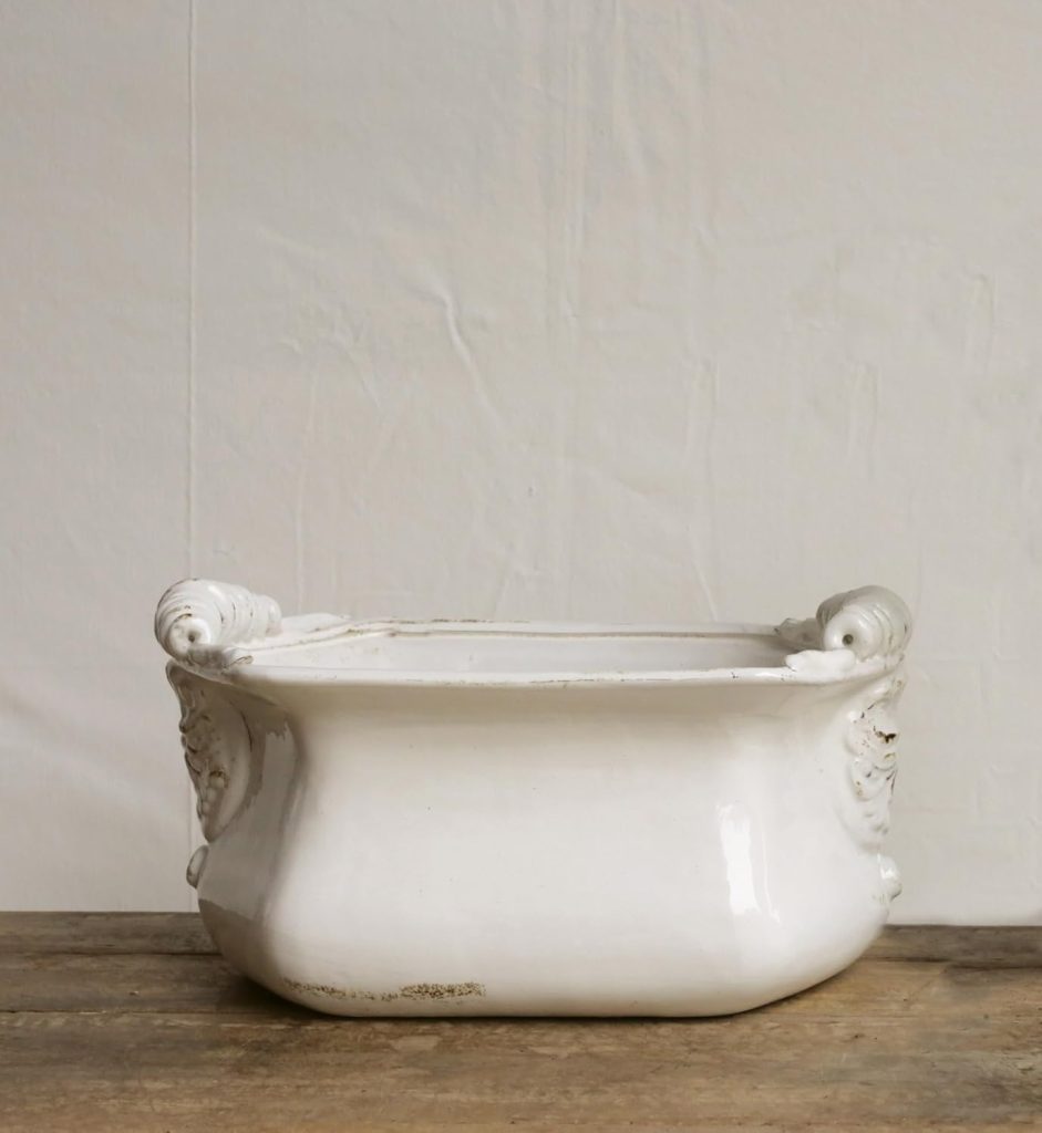 White ceramic chamber pot with ornate handles sits on a wooden surface against a plain, light-colored wall, evoking the charm of rustic outdoor fall decor.