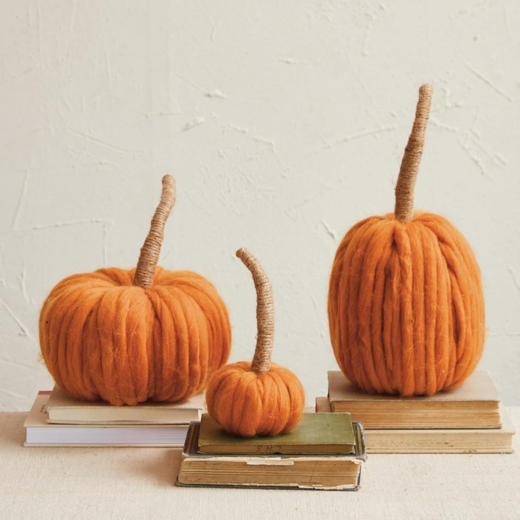 Three orange yarn pumpkins with wrapped stems from A Home to Share are displayed on stacked books against a neutral textured wall.