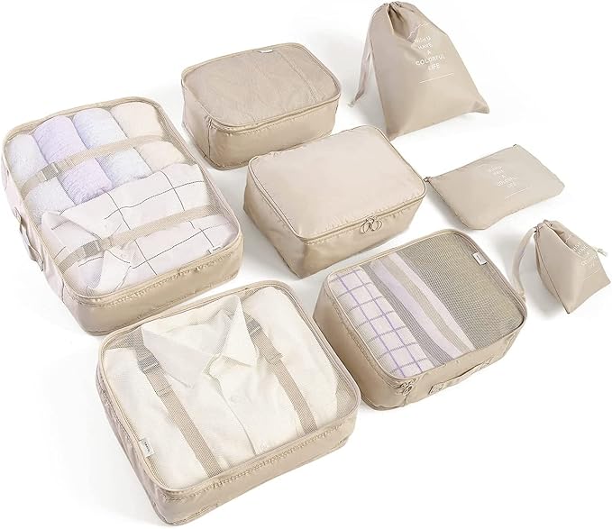 A set of beige travel packing cubes and bags are displayed, containing folded clothes and various items. The set includes different sizes of cubes, from large to small, with transparent mesh tops.