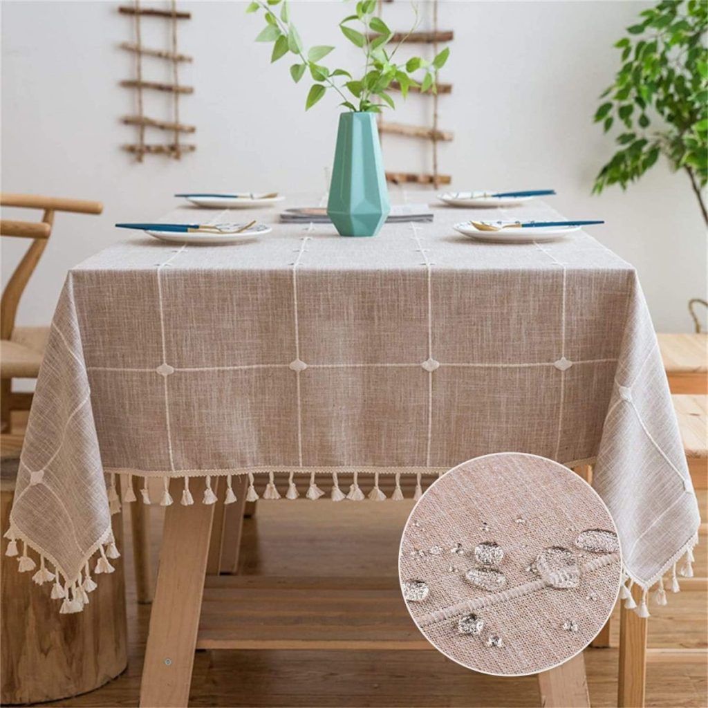 A dining table is covered with a light brown checkered tablecloth featuring tassels and set with plates and cutlery. A green vase with leafy branches is placed at the center. Inset shows water droplets on the cloth.