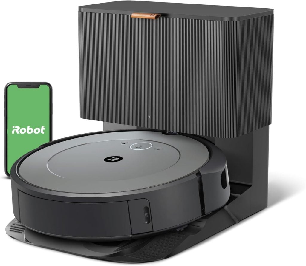 A robotic vacuum cleaner is docked at its charging station, with a smartphone displaying the iRobot app nearby.