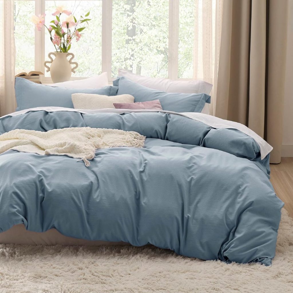 A neatly made bed with light blue bedding and several pillows near a large window. A white vase with pink flowers is on the window sill, and a fluffy white rug is on the floor.