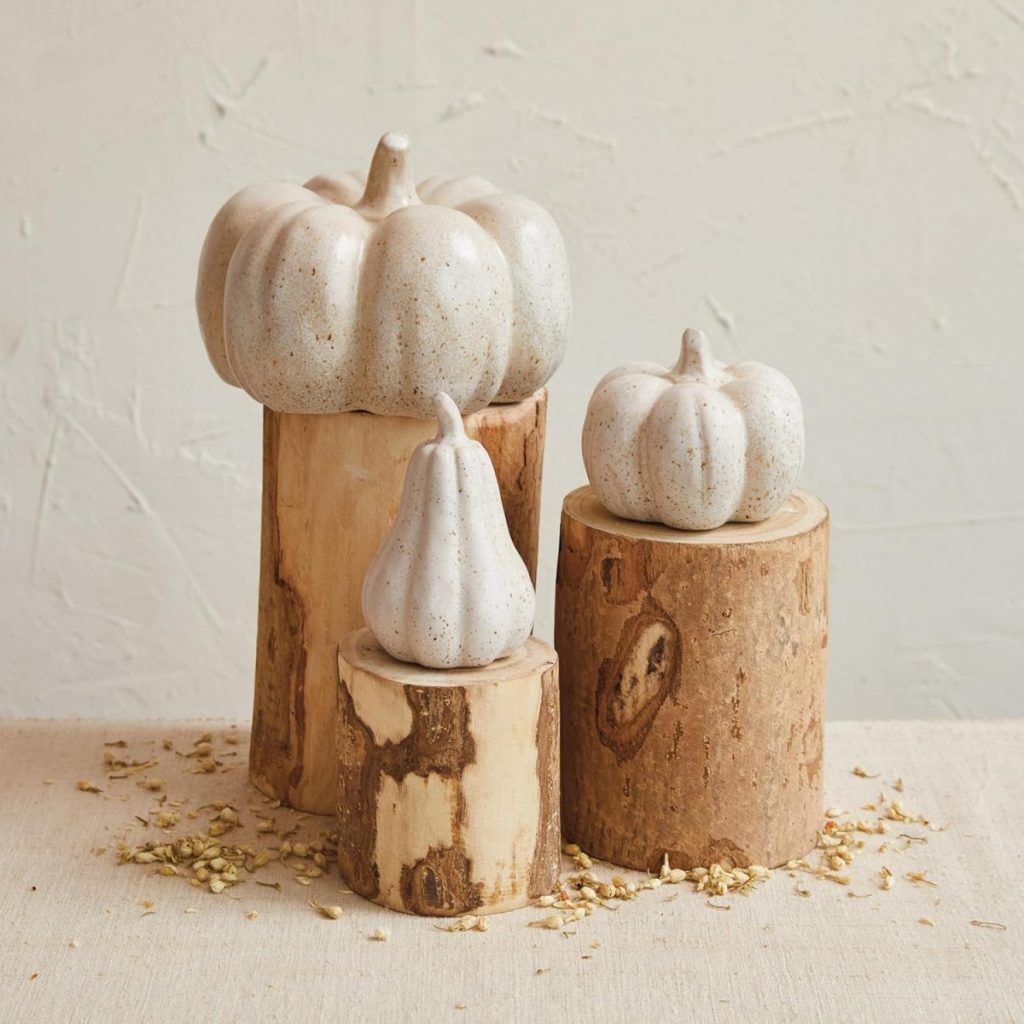 Three white ceramic gourds from A Home to Share are displayed on cut wooden logs of varying heights, set on a neutral fabric surface with scattered dried flower petals.
