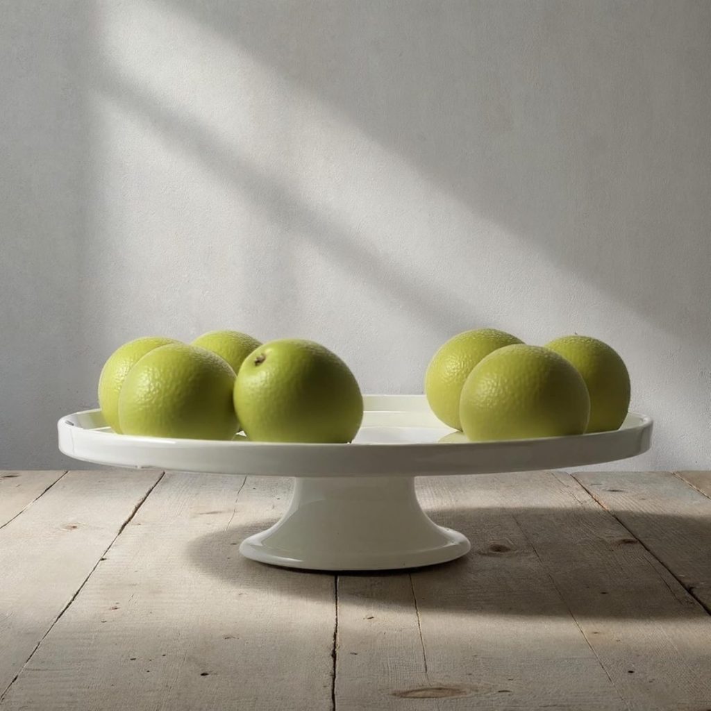 Seven green apples arranged on a white pedestal cake stand, placed on a wooden table with sunlight casting shadows, evoke the cozy feel of autumn—perfect alongside a warm pumpkin spice latte.