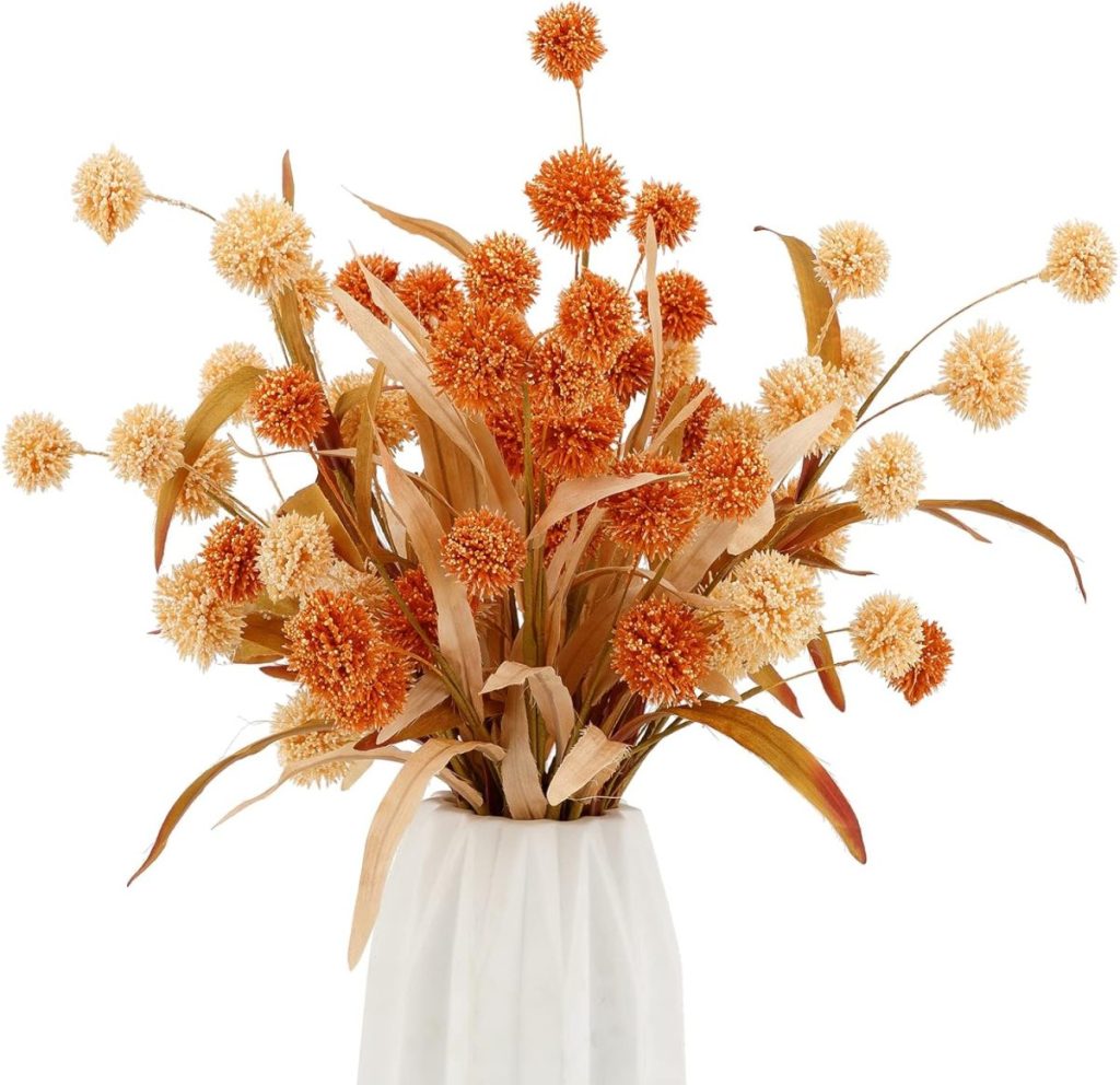A white vase with an arrangement of orange and cream dried billy button flowers and long brown leaves, perfect for fall flower arrangements, set against a white background.
