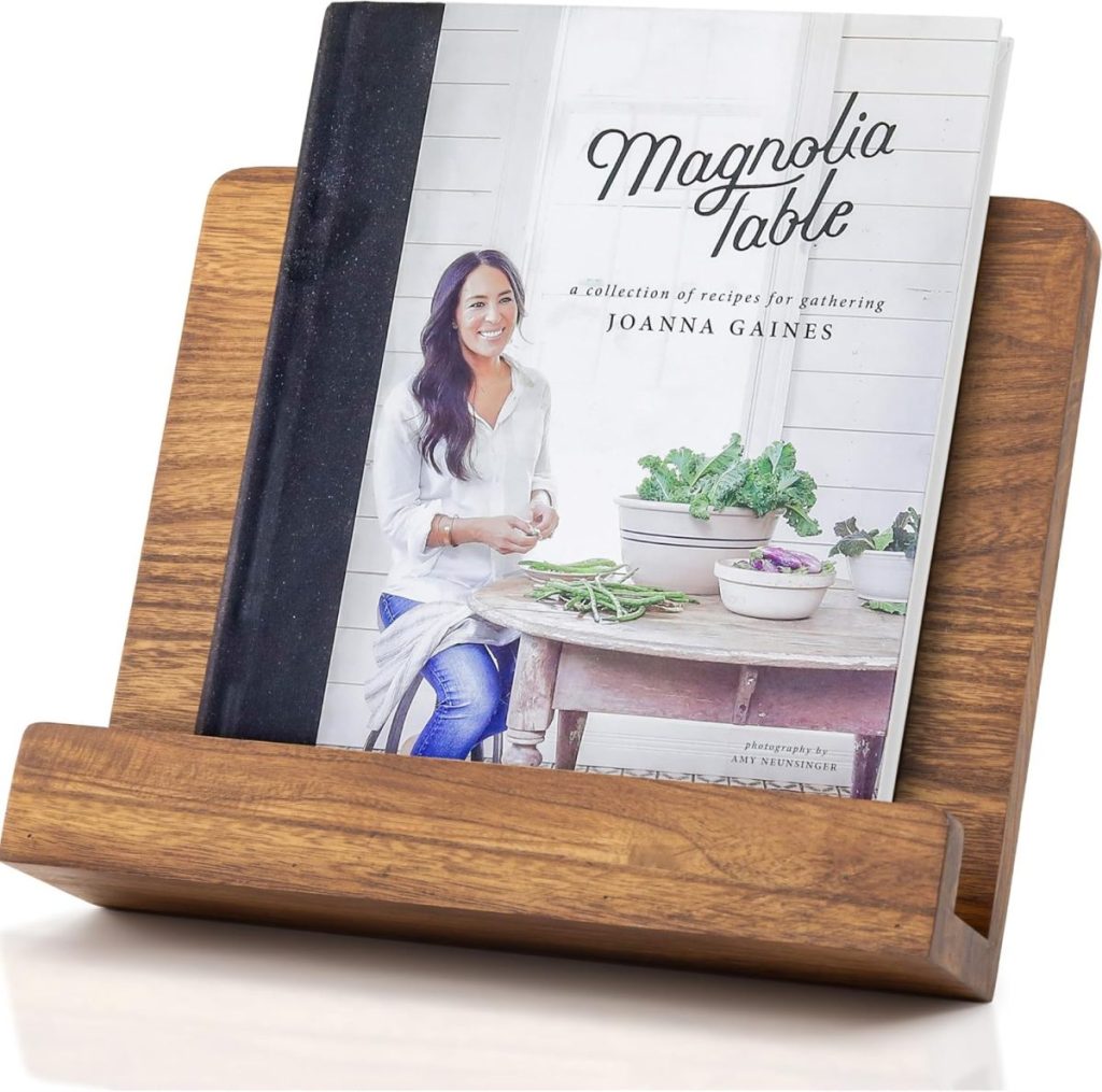 A cookbook titled "Magnolia Table" by Joanna Gaines is displayed on a wooden book stand, creating a warm scene reminiscent of A Home to Share.