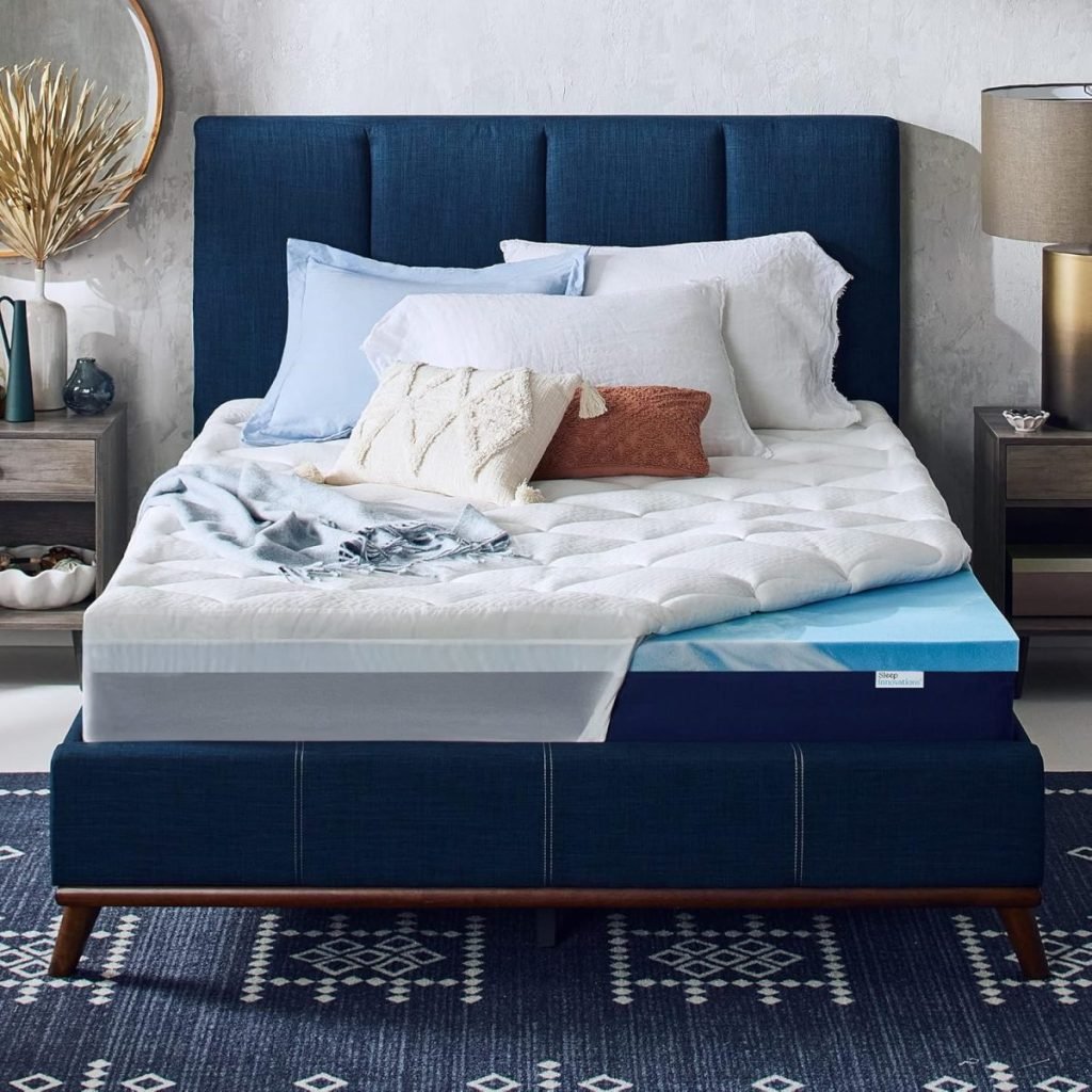 A neatly made bed with a navy blue headboard and various pillows. The mattress has a cutaway section showing multiple layers. A bedside table with a lamp and decor is on the right side.