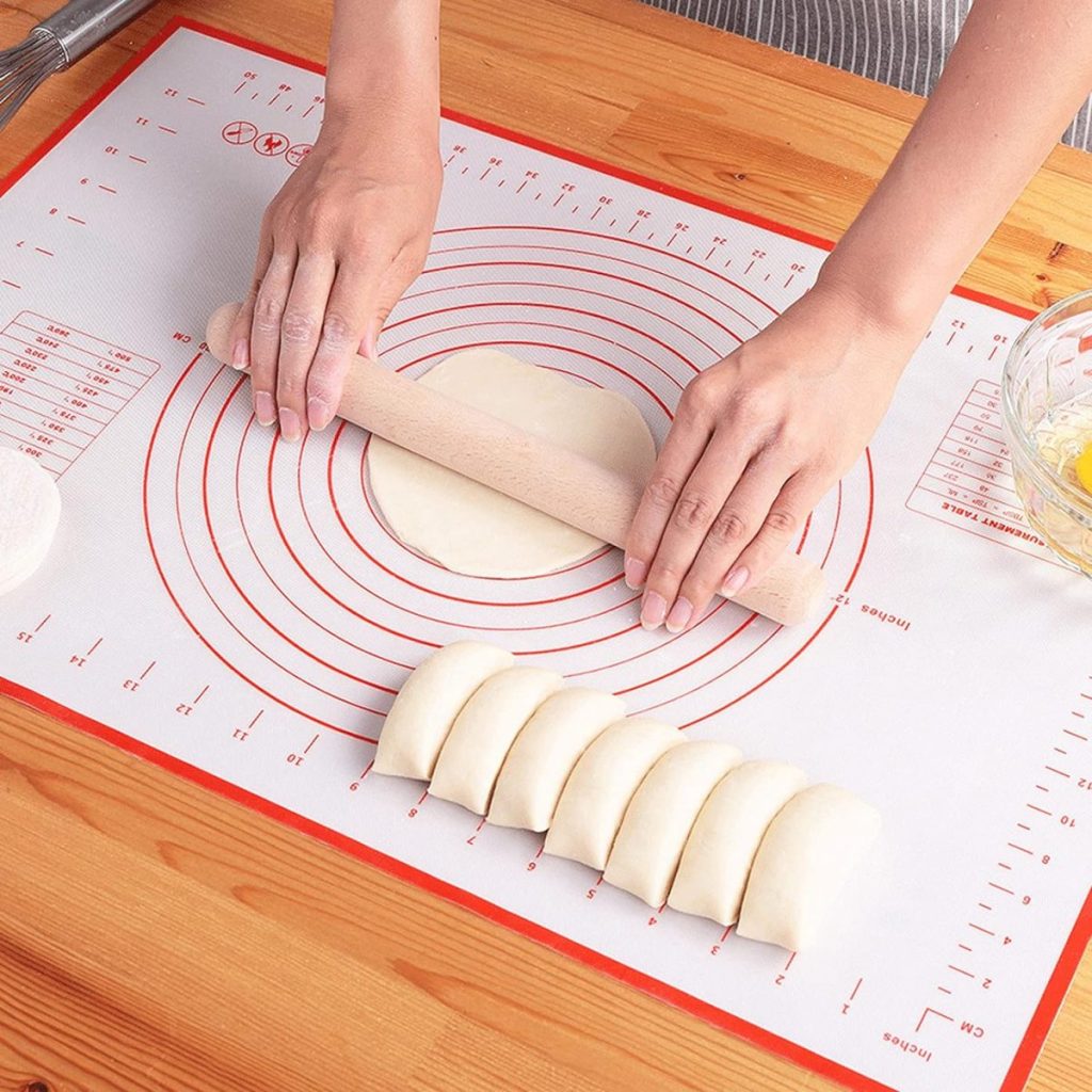 Person rolling out dough with a rolling pin on a marked silicone baking mat, with rolled and sliced dough pieces nearby on a wooden surface.