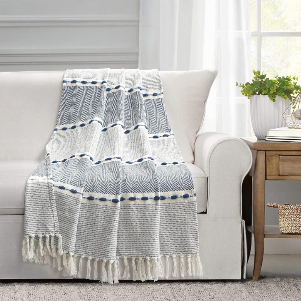 A white couch with a blue and white woven throw blanket draped over the backrest. There's a wooden side table with books, a potted plant, and a woven basket underneath by the couch.