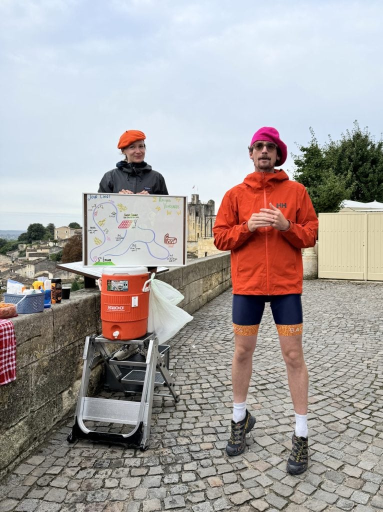 Two people stand outdoors on cobblestone, one in an orange jacket and pink hat, the other in gray with an orange beret, next to a map display and a large orange drink cooler.