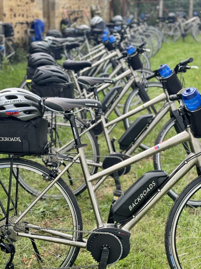 A row of parked bicycles with "Backroads" written on them, equipped with helmets and water bottles, lined up on grass.