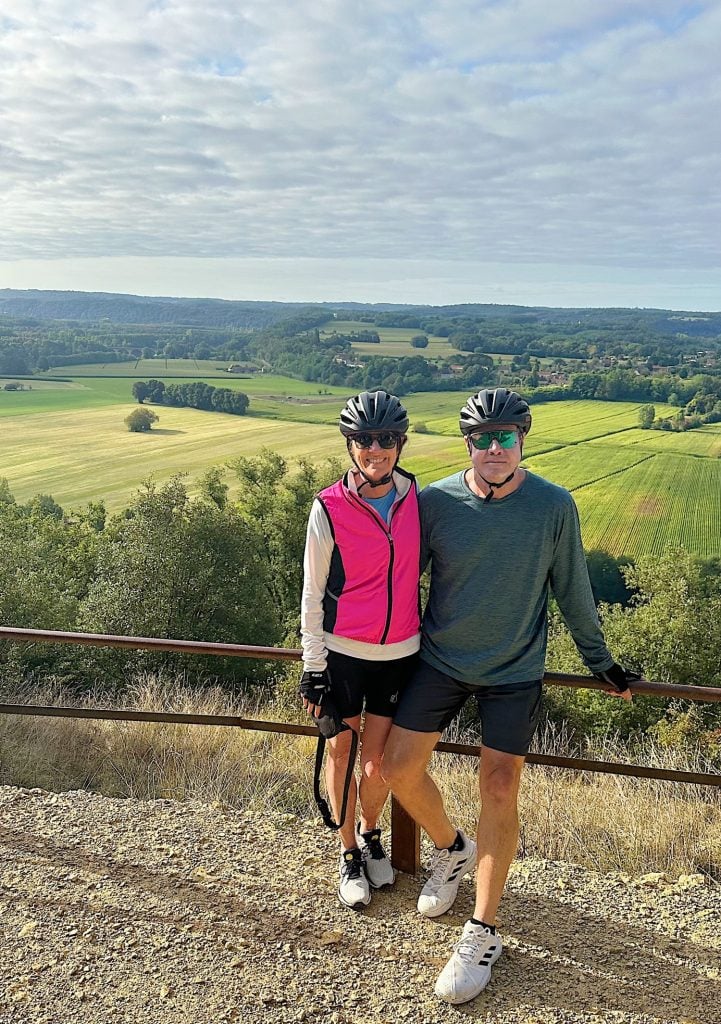 A man and woman wearing cycling gear, including helmets and gloves, stand together on a gravel road overlooking a green and yellow landscape with trees and a cloudy sky.