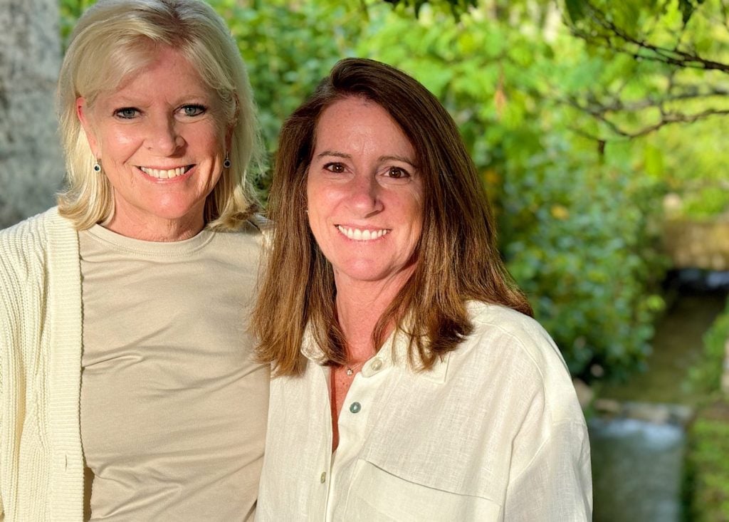 Two women stand outdoors, smiling at the camera, with green foliage and a stream in the background.