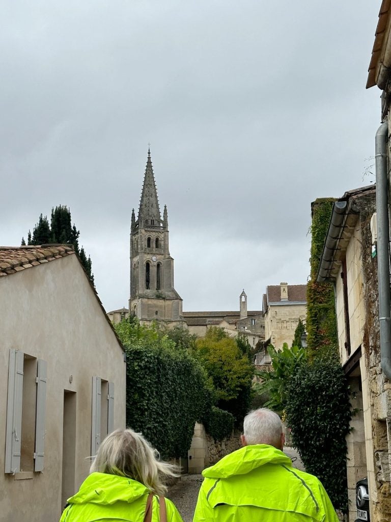 Two people in bright yellow jackets walk down a narrow pathway towards a historic stone church with a tall spire on a cloudy day. The path is lined with buildings and lush greenery.