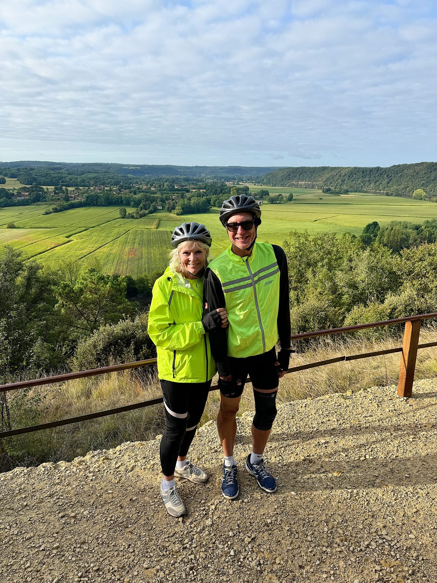 Two cyclists wearing helmets and bright jackets stand next to each other on a gravel path with expansive, green countryside in the background.