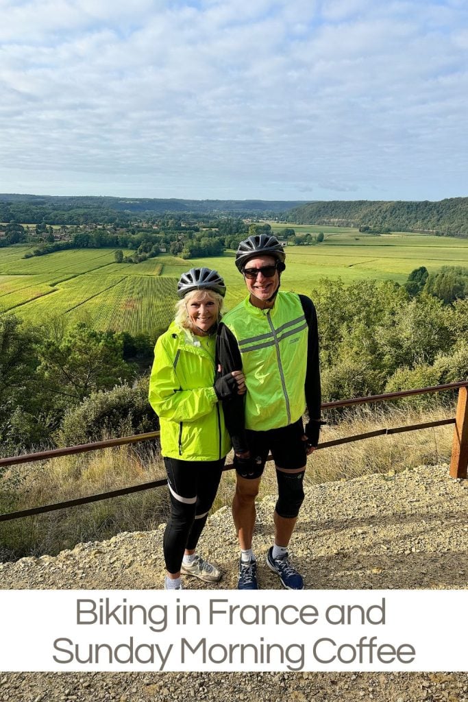 Two cyclists in bright green jackets stand together smiling, with a scenic view of fields and hills in the background. Text below reads, "Biking in France and Sunday Morning Coffee.