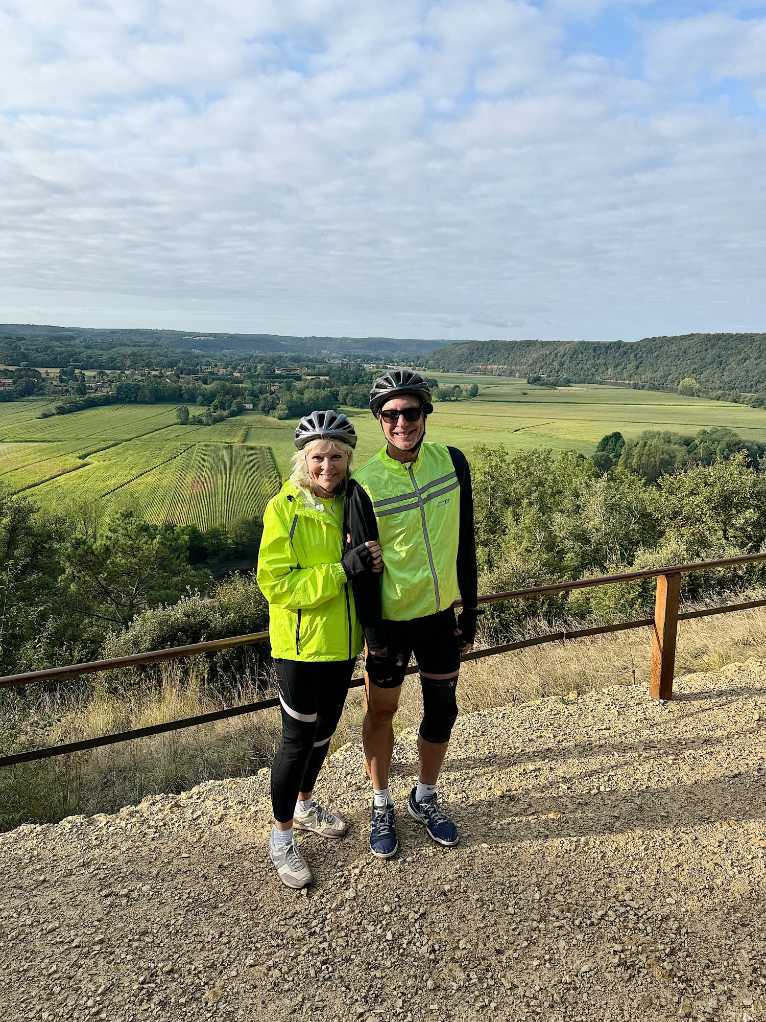 Two cyclists in bright green jackets and helmets stand on a cliffside path overlooking a vast, green landscape with rolling hills and a cloudy sky.