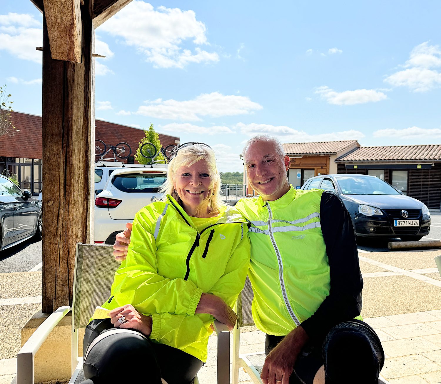 Two people wearing bright yellow jackets sit side by side on a bench outdoors. They are smiling and have bicycles mounted on a car in the background.