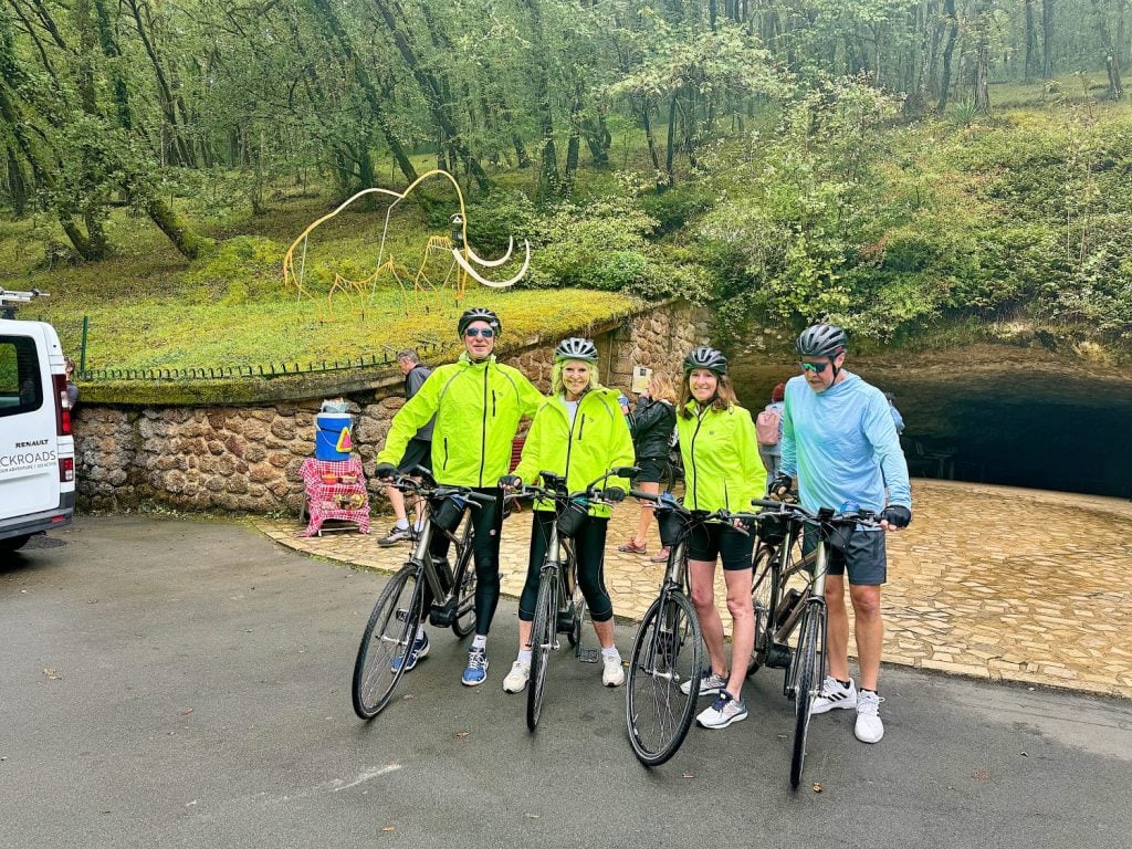 Four cyclists in green and blue jackets, wearing helmets, stand with their bikes in front of a cave entrance surrounded by greenery and a mammoth sculpture. A white van is parked nearby.