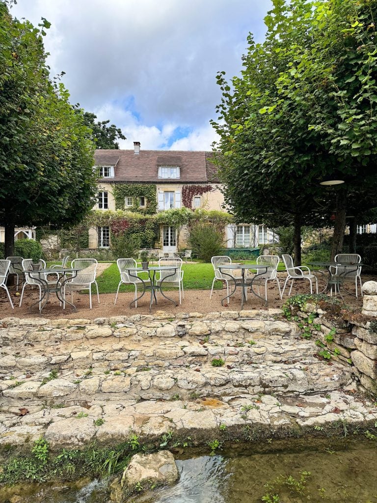 Outdoor seating area with empty metal chairs and tables on a stone patio, surrounded by lush greenery and a quaint house in the background.