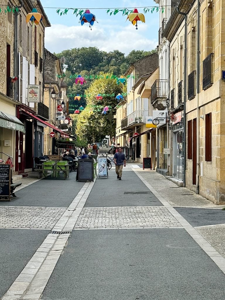 A pedestrian walks down a cobblestone street lined with outdoor cafes and shops. Umbrellas hang overhead as decorations, and the street extends toward a green, wooded area in the background.