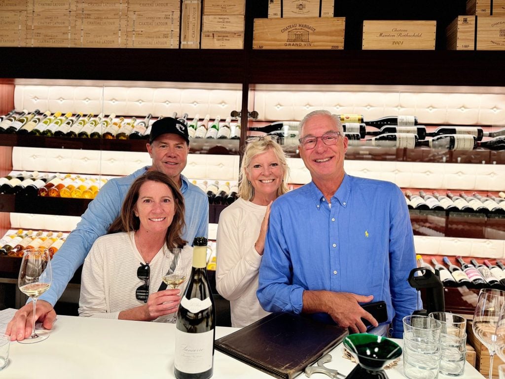 Four people are standing in front of a wall of wine bottles at a wine tasting. Three of them are holding glasses of wine and smiling at the camera.