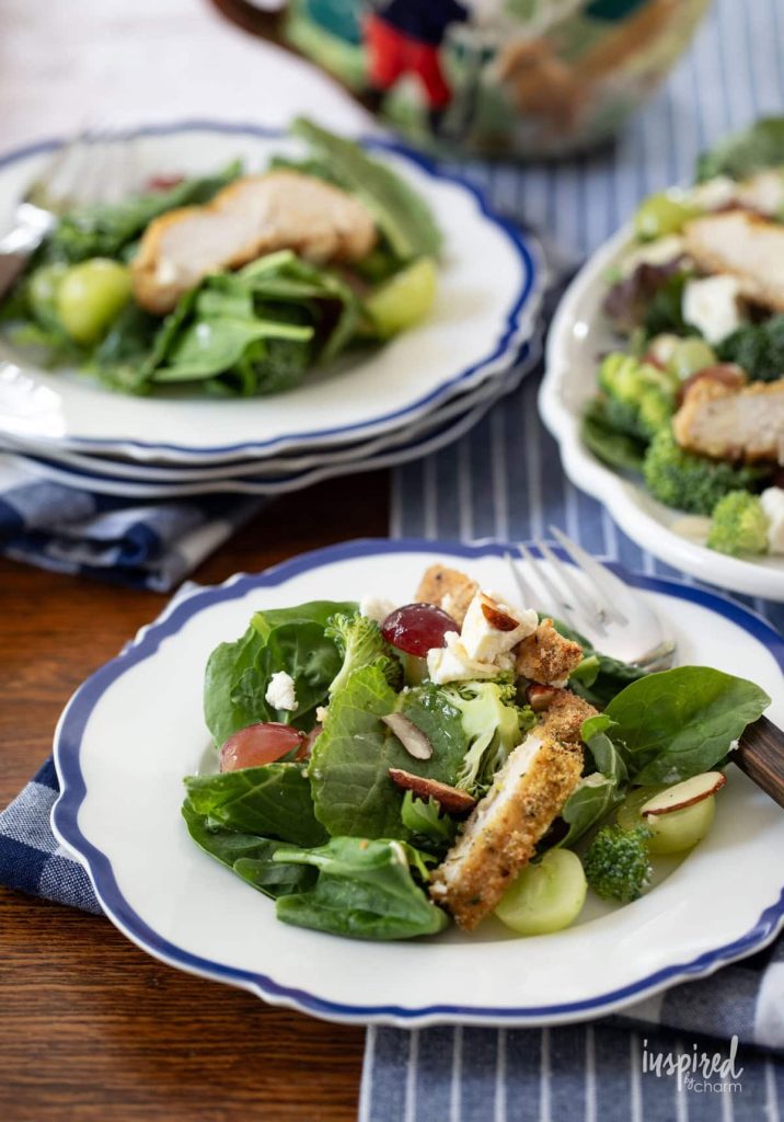 Plate of fresh green salad with mixed greens, grapes, broccoli, almonds, and pieces of breaded chicken. Fork on the side, another similar plate and a salad bowl in the background.