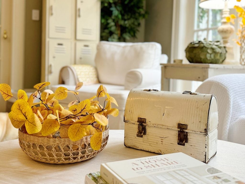 A white room with lockers and white furniture. A table features a white vintage lunchbox, a wicker basket with yellow leaves, and a book titled "A Home to Share.