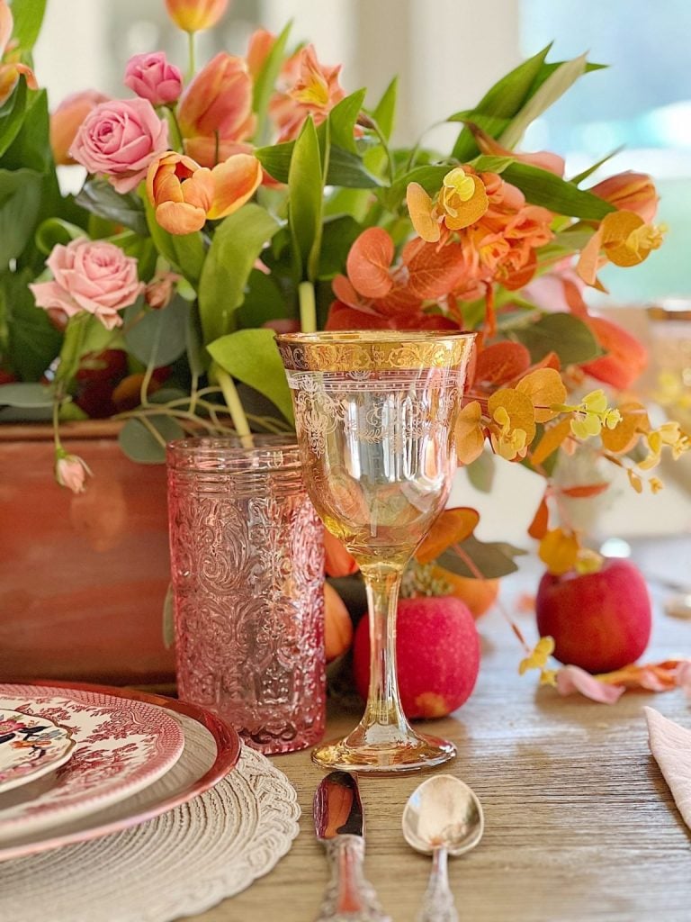 A decorated dining table with a floral centerpiece, featuring pink and orange flowers. There is an ornate wine glass, a textured tumbler, a patterned plate, and silver cutlery. Apples are scattered around.