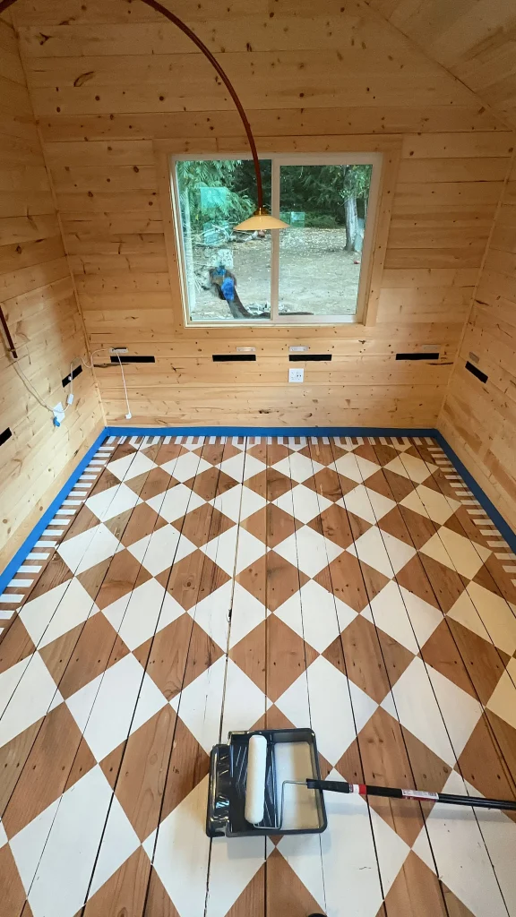 A wooden room with a painted diamond-patterned floor, a paint roller on a tray in the foreground, and a window showing trees outside.