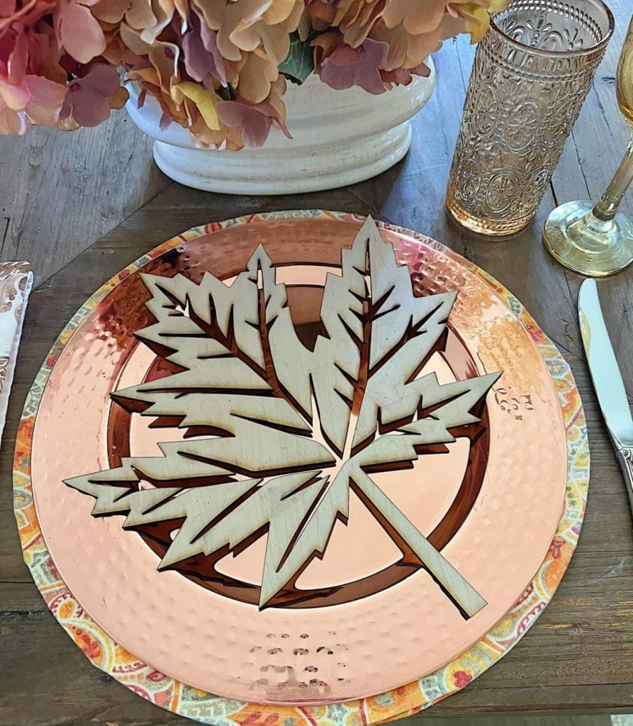 A table setting with a large metal maple leaf decoration placed on an orange charger plate, surrounded by a knife, fork, patterned napkin, and gold-toned drinkware.