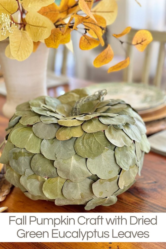 A pumpkin decorated with dried green eucalyptus leaves sits on a wooden table. Yellow leaves in a vase are in the background. Text below reads "Fall Pumpkin Craft with Dried Green Eucalyptus Leaves.