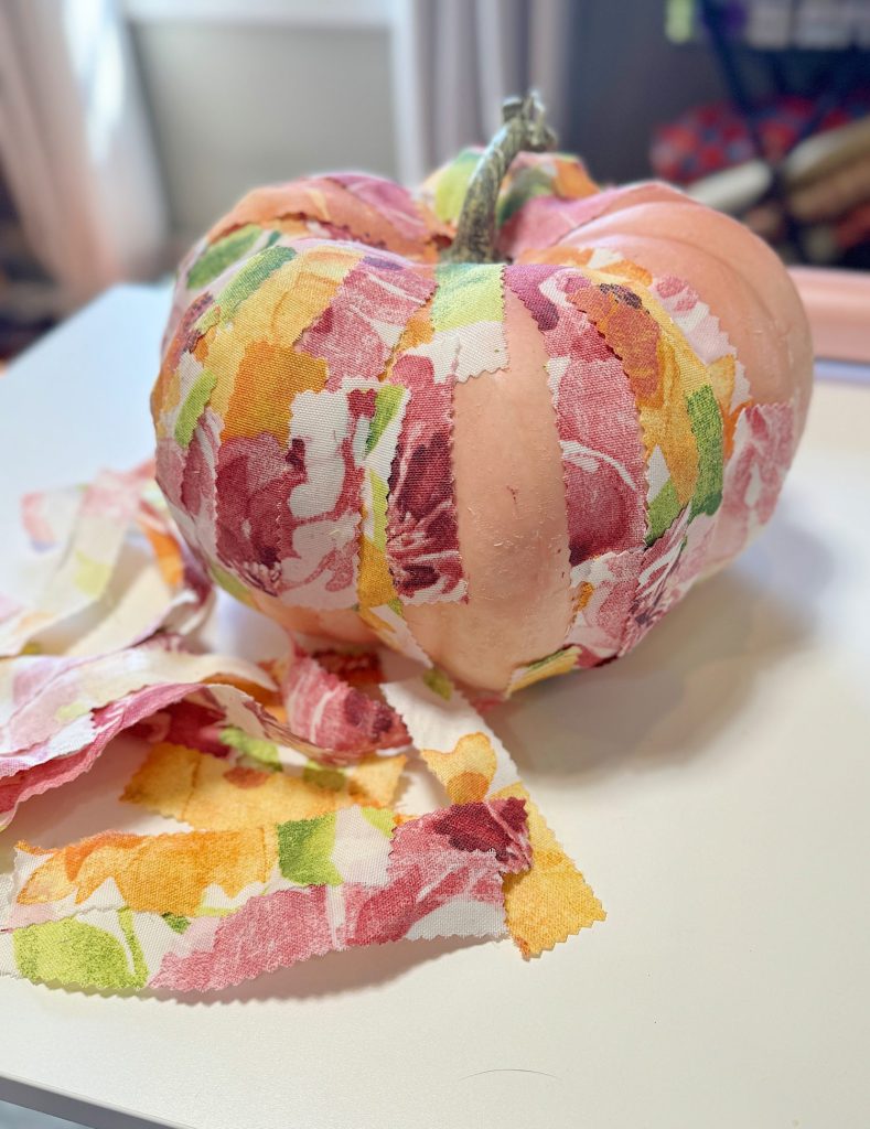 A pumpkin is decorated with colorful fabric strips, sitting on a white surface, with additional fabric strips nearby.