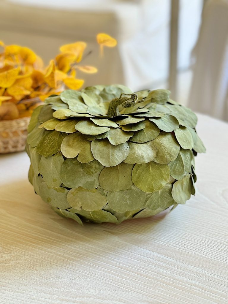 A decorative pumpkin covered with green leaves sits on a light wooden table next to a basket of yellow leaves.