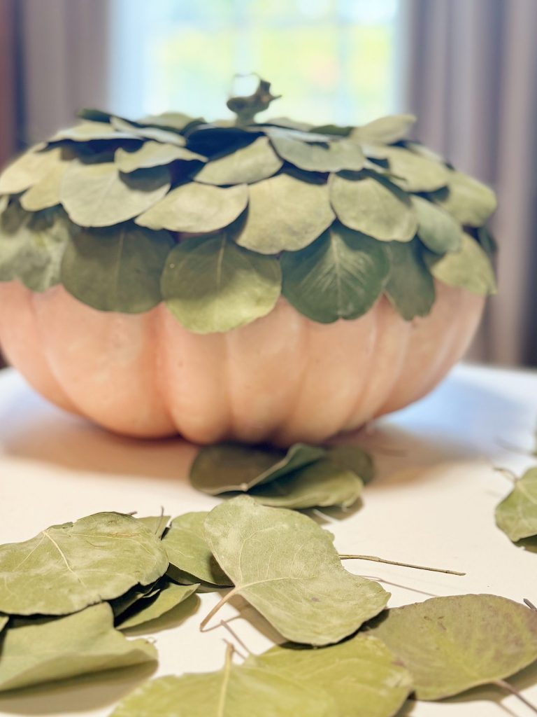 A large pumpkin with a top covered in green leaves sits on a table, surrounded by additional leaves.