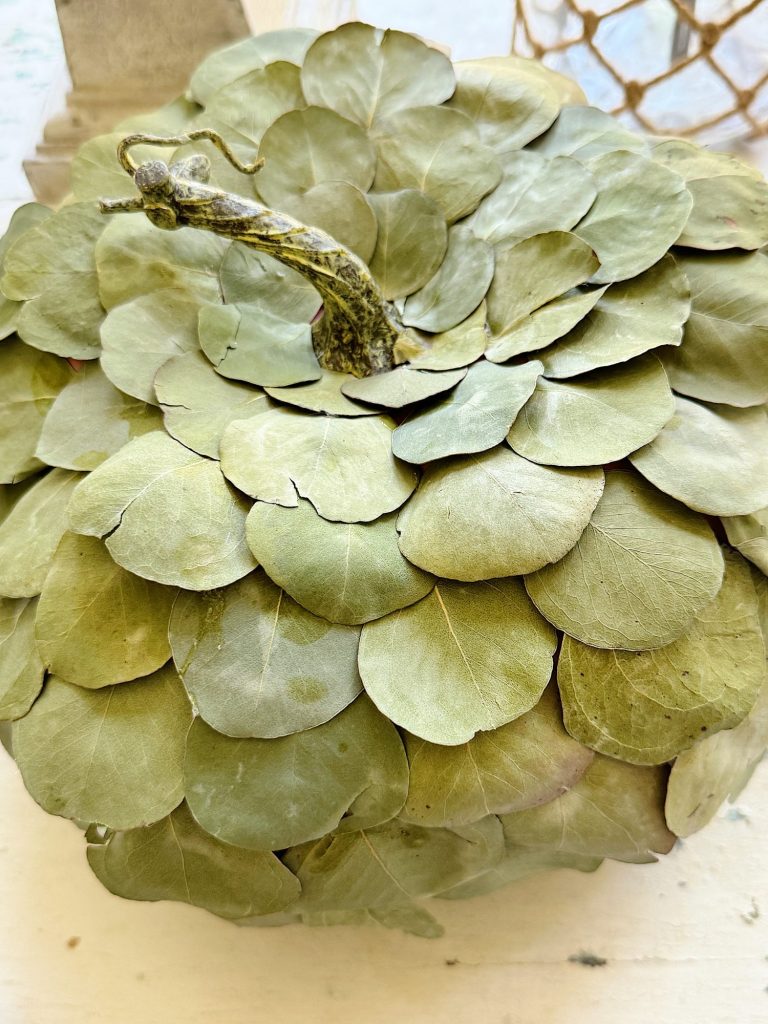 A spherical object covered entirely with overlapping dried green leaves, with a protruding stem-like structure at the top.